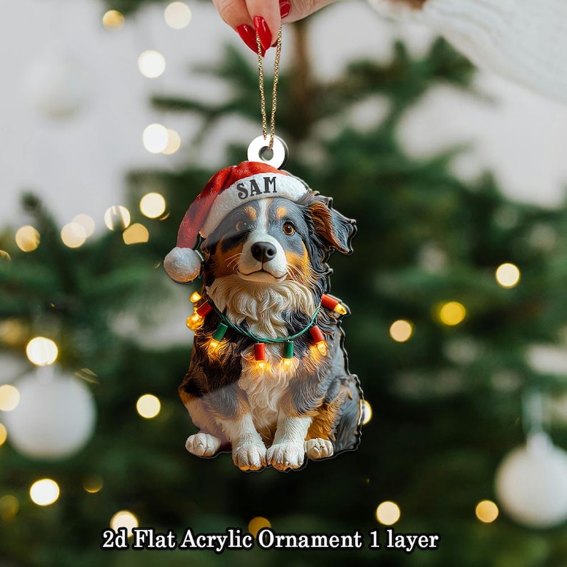 May include: A 2D flat acrylic Christmas ornament featuring a dog wearing a Santa hat with the name "SAM" and a string of lights. The dog is black, brown, and white. The ornament is hanging in front of a blurred Christmas tree.