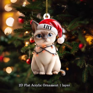 May include: A 2D flat acrylic Christmas ornament featuring a Siamese cat wearing a Santa hat with the name "LILY". The cat has blue eyes and is adorned with a string of lights. The ornament hangs in front of a Christmas tree.