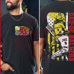 Op de afbeelding: Zwart T-shirt met een rood-wit-blauw Amerikaans vlagontwerp. De vlag toont twee honkballers. De achterkant van het shirt heeft de tekst "Dad of Ballers" met een honkballer in een rood-wit-blauw uniform. De naam van de speler is "Joe" en zijn nummer is "8".