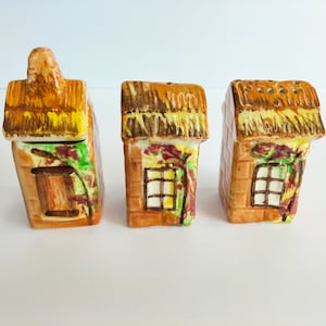 May include: Three ceramic salt and pepper shakers shaped like cottages with thatched roofs and painted details. The cottages are brown with yellow roofs and green vines.