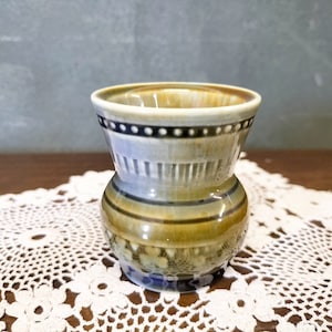 May include: A small, decorative ceramic vase with a flared top and a bulbous base. The vase features a color palette of tan, blue, and brown, with a band of dark blue dots near the rim. The vase is approximately 4 inches tall.