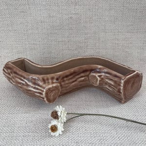 May include: A brown ceramic log-shaped planter with a wavy design. The planter has a textured surface and is decorated with a small sprig of white flowers.