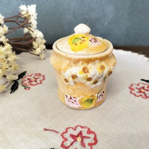 May include: A small ceramic jar with a lid, decorated with yellow and pink floral designs. The jar is a light beige colour and sits on a floral embroidered cloth. The lid has a small, white knob.