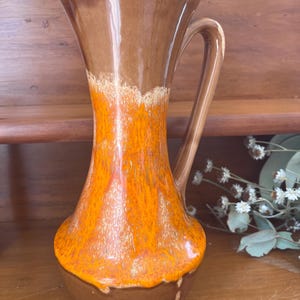 May include: A vintage ceramic jug with a handle. The jug has a brown base that blends into a bright orange and brown upper section. The rim is a lighter brown. The jug is likely for decorative use.