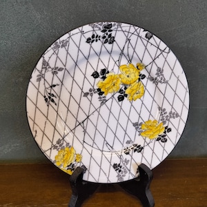 May include: A white ceramic plate with a black and grey trellis pattern and yellow roses. The plate has a black rim.