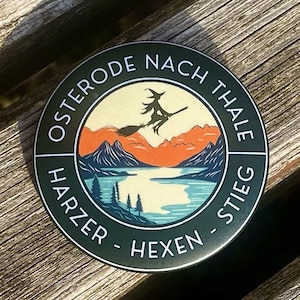 Könnte beinhalten: Ein runder, grüner Aufkleber mit dem Text "Osterode nach Thale Harzer Hexen-Stieg" in einem Kreis um eine Grafik einer Hexe, die auf einem Besen über einem See und Bergen fliegt.
