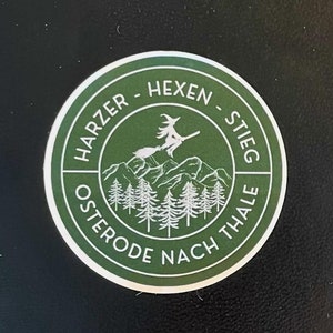 Könnte beinhalten: Ein schwarzes Notizbuch mit einem grünen runden Aufkleber, auf dem "Harzer Hexen-Stieg Osterode nach Thale" steht und eine Hexe auf einem Besen über einem Gebirge fliegt.