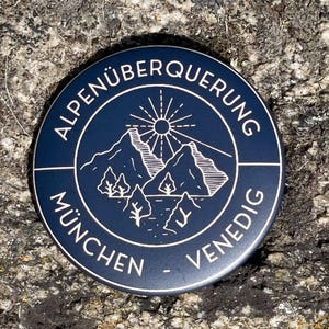 Op de afbeelding: Een donkerblauwe ronde knoop met witte tekst en een bergketenafbeelding. De tekst luidt "Alpenüberquerung München - Venedig". De afbeelding toont bergen, bomen en een zonnestraal. De knoop ligt op een getextureerd, grijs stenen oppervlak.