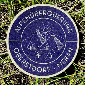 Könnte beinhalten: Ein runder, blau-weißer Aufkleber mit dem Text "ALPENÜBERQUERUNG OBERSTDORF - MERAN", der ein Berg- und Sonnengrafik umrandet. Der Aufkleber liegt auf einem Hintergrund aus grünem Gras.