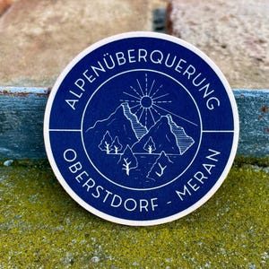 Op de afbeelding: Een ronde, marineblauwe sticker met witte tekst en een berg- en zongrafiek. De tekst luidt "ALPENÜBERQUERUNG OBERSTDORF - MERAN" in een cirkelvormig patroon. De sticker ligt op een gestructureerd, mosachtig oppervlak.