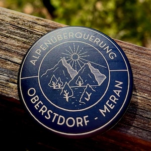 Könnte beinhalten: Ein runder, dunkelblauer Anstecker mit einer weißen Linienzeichnung von Bergen, Bäumen und einer Sonne. Der Text ALPENÜBERQUERUNG ist oben bogenförmig angeordnet, während OBERSTDORF - MERAN am unteren Rand des runden Designs steht.