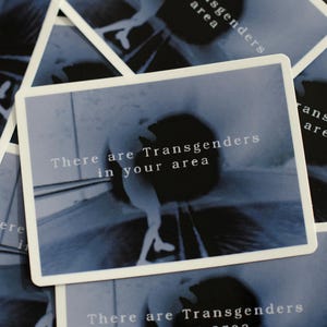 Może przedstawiać: Kilka kart z białymi obramowaniami leży na ciemnej powierzchni. Każda karta zawiera niebieski obraz i tekst "There are Transgenders in your area" w białym kolorze. Karty są lekko nachodzące na siebie.