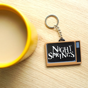 Op de afbeelding: Een gele mok gevuld met koffie of thee staat naast een sleutelhanger met een bruin en zwart televisie-vormig sieraad. Het sieraad heeft de woorden "Night Springs" in wit op het scherm.