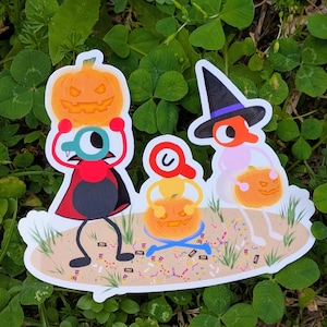 Op de afbeelding: Een gestanste sticker met drie stripfiguren die Halloween vieren. Eén figuur draagt een vampiercape en houdt een pompoen vast, een ander zit met een pompoen, en de derde draagt een heksenhoed en houdt ook een pompoen vast.