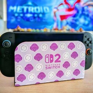 May include: A black Nintendo Switch console with a purple and white patterned cover. The cover features a repeating design of stylized flowers and the words "Nintendo Switch". A television screen is visible in the background.