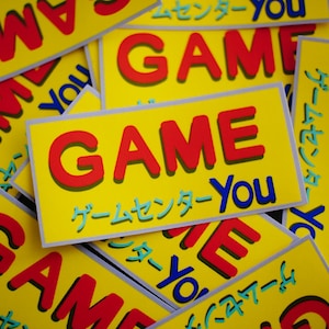 Könnte beinhalten: Gelbe rechteckige Aufkleber mit dem Wort "GAME" in Rot, weiß umrandet, und dem Wort "You" in Blau. Japanische Schriftzeichen sind ebenfalls vorhanden. Die Aufkleber sind verstreut.