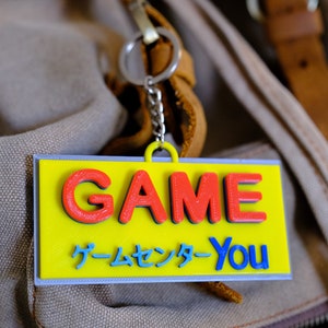 Può includere: Un portachiavi giallo e blu con la parola "GAME" in lettere rosse e il testo "ゲームセンター-You" in lettere blu.