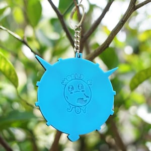 May include: A bright blue keychain with a circular design. The keychain features a cartoon globe with a smiling face and the words "WORLD & JOY" in white. The keychain is hanging from a silver chain against a blurred green background.