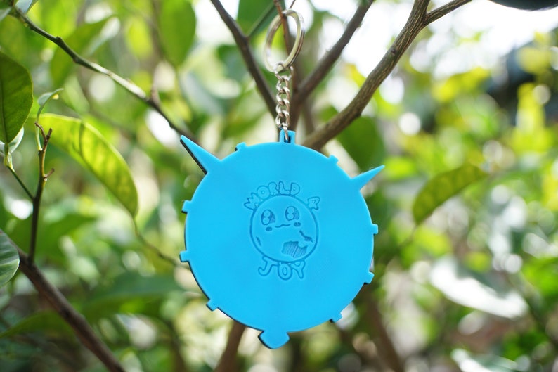 May include: A bright blue keychain with a circular design. The keychain features a cartoon globe with a smiling face and the words "WORLD & JOY" in white. The keychain is hanging from a silver chain against a blurred green background.
