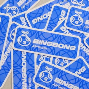 Puede incluir: Un conjunto de pegatinas rectangulares azules con esquinas redondeadas. Cada pegatina presenta el texto "BINGBONG Airlines" en blanco, un personaje de dibujos animados y un diseño de globo terráqueo. Las pegatinas están dispersas sobre una superficie clara.