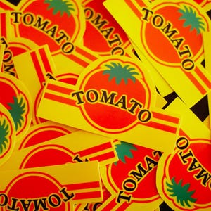 Può includere: Una collezione di adesivi gialli con una grafica di pomodoro rosso e la scritta "TOMATO". L'illustrazione del pomodoro ha foglie verdi. Gli adesivi hanno strisce rosse e arancioni.