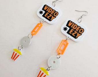 May include: A pair of dangle earrings featuring a white square charm with the black and orange text VIDEO EZY, a small orange rectangular cassette tape charm, a silver circular disc, and a small white and orange popcorn bucket charm.