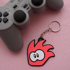 Może przedstawiać: Różowo-biały brelok do kluczy z postacią z kreskówki o głowie w kształcie płomienia i dużymi oczami. Brelok jest przymocowany do srebrnego łańcuszka i srebrnego kółka.
