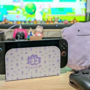 Könnte beinhalten: Eine schwarze Nintendo Switch-Konsole mit einem lila Dock mit einem Ditto-Design und dem Nintendo Switch-Logo. Ein Ditto-Plüschtier und ein schwarzer Controller sind ebenfalls zu sehen. Der Hintergrund umfasst einen Fernseher und verschiedene dekorative Gegenstände.
