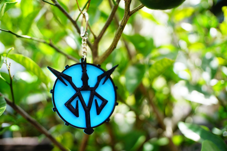 May include: A round keychain with a light blue center and a black emblem. The emblem features a stylized design with arrows and a central symbol. The keychain is attached to a silver chain and ring, set against a blurred green background.