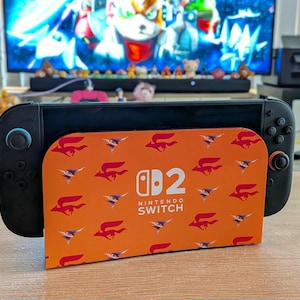 Puede incluir: Una consola Nintendo Switch negra con detalles en azul y naranja, apoyada en un soporte. El soporte es naranja y presenta un patrón repetido de un pájaro estilizado y el texto "Nintendo Switch". La consola está sobre una superficie de madera.