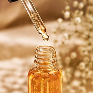 K&ouml;nnte beinhalten: Eine klare Glasflasche gef&uuml;llt mit einer gold-bernsteinfarbenen Fl&uuml;ssigkeit, ausgestattet mit einer schwarzen Pipettenkappe. Eine Glaspipette schwebt &uuml;ber der offenen Flasche, wobei ein Tropfen der goldenen Fl&uuml;ssigkeit an der Spitze h&auml;ngt.