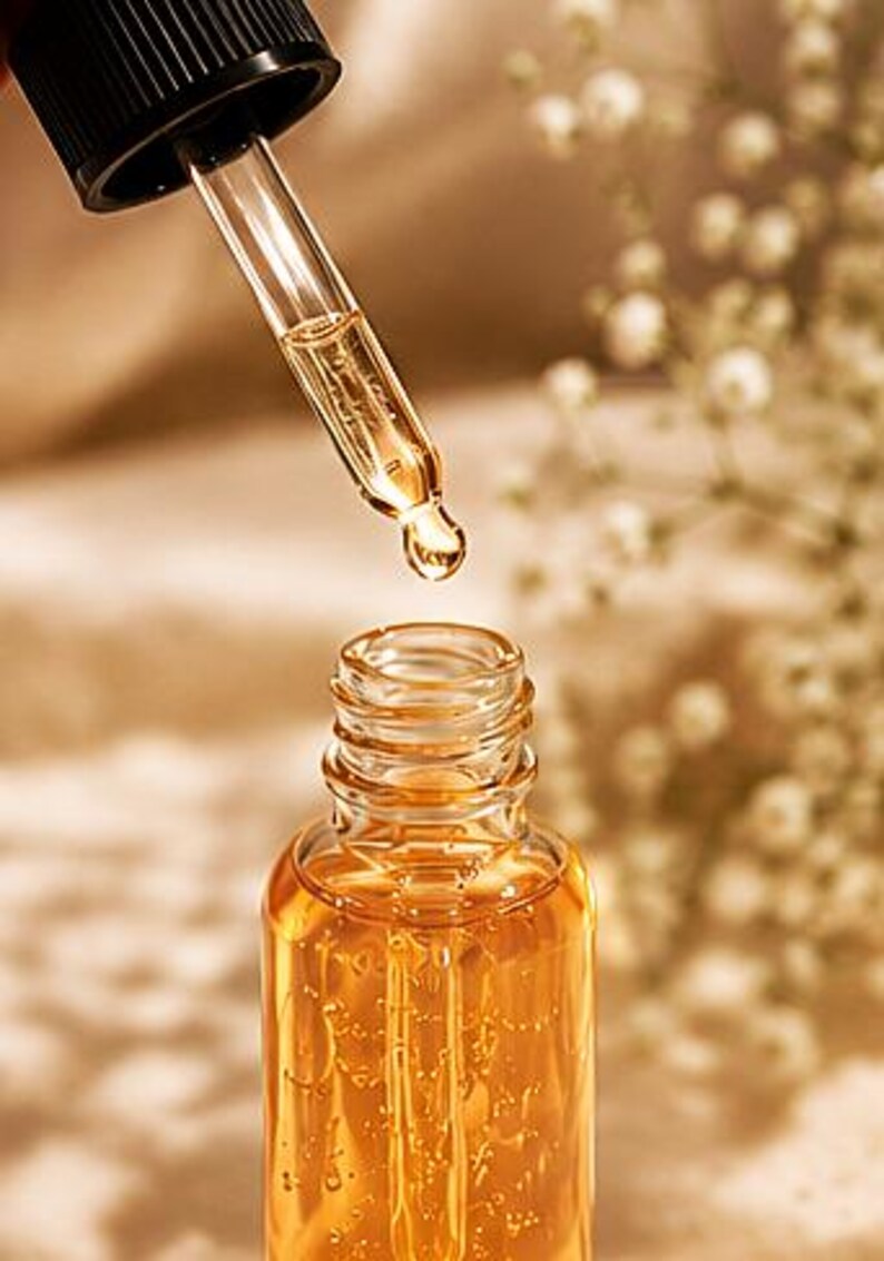 K&ouml;nnte beinhalten: Eine klare Glasflasche gef&uuml;llt mit einer gold-bernsteinfarbenen Fl&uuml;ssigkeit, ausgestattet mit einer schwarzen Pipettenkappe. Eine Glaspipette schwebt &uuml;ber der offenen Flasche, wobei ein Tropfen der goldenen Fl&uuml;ssigkeit an der Spitze h&auml;ngt.