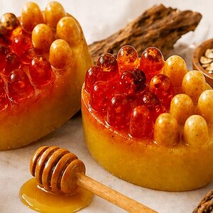 May include: Two handmade soaps with a unique design, featuring a yellow base and topped with amber-colored, rounded elements. A wooden honey dipper rests on a small puddle of honey. Oats and turmeric are in small wooden bowls.