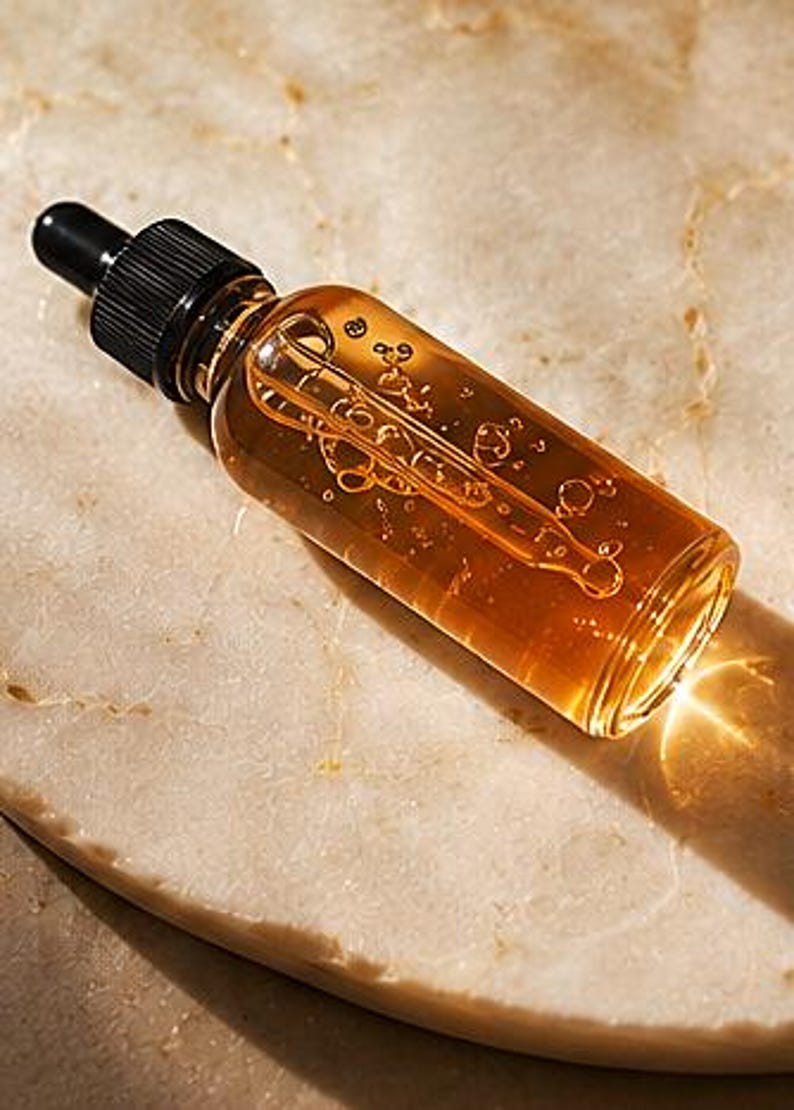 K&ouml;nnte beinhalten: Eine klare Glastropfflasche gef&uuml;llt mit einer gold-bernsteinfarbenen Fl&uuml;ssigkeit. Die Flasche hat einen schwarzen, gerippten Schraubverschluss mit einem schwarzen Gummiball. Im Inneren ist eine Glaspipette sichtbar, die in dem durchscheinenden &Ouml;l liegt.