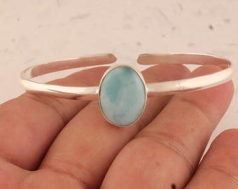 May include: A silver cuff bracelet featuring a smooth, polished metal band and a central oval-shaped larimar gemstone. The stone displays a marbled pattern of light blue and white tones, securely set within a simple, minimalist silver bezel setting.