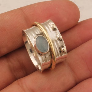 Puede incluir: Un anillo giratorio de plata y oro con una piedra ovalada azul claro. El anillo tiene una banda texturizada y tres cuentas doradas.
