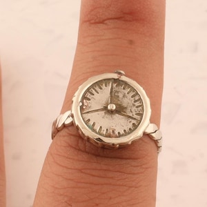 Könnte beinhalten: Ein silberner Ring mit einem Zifferblatt-Design. Das Zifferblatt des Rings hat ein detailliertes Zifferblatt mit Zeigern, die die Uhrzeit anzeigen. Das Band hat ein geflochtenes Design. Der Ring wird an einem Finger getragen.