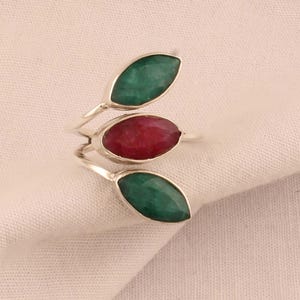 Op de afbeelding: Een zilveren ring met drie edelstenen. De middelste steen is dieprood, en de twee stenen aan weerszijden zijn felgroen.