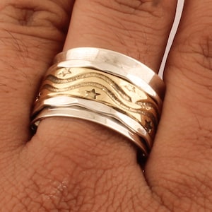 May include: A silver and gold spinning ring with a wavy design and stars. The ring is worn on a finger.