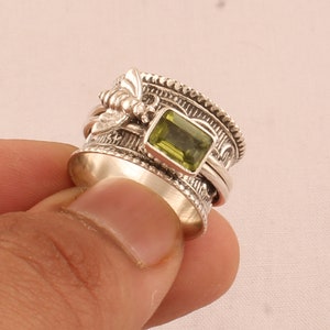 May include: A silver ring featuring a green gemstone and a bee design. The ring is made of two bands that spin around each other.