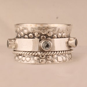 Puede incluir: Un anillo giratorio de plata con una banda texturizada y tres piedras preciosas transparentes. El anillo está diseñado para girar libremente en el dedo.