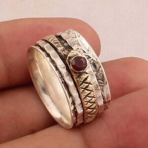 Puede incluir: Un anillo giratorio de plata y oro con una piedra preciosa roja. El anillo tiene un diseño texturizado y está hecho de múltiples bandas.