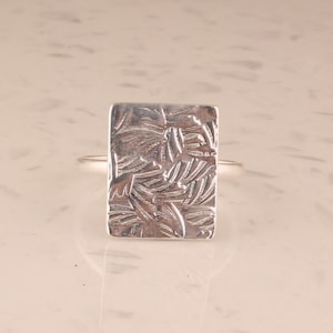 以下が含まれることがあります： テクスチャ加工された長方形の面を持つ銀の指輪。テクスチャは葉のパターンに似ています。