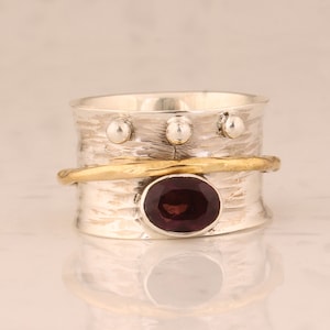 May include: A silver-tone ring with a textured band, featuring a gold-tone band and a dark red oval gemstone. The ring has three small silver-tone spheres on the top. The ring is designed to spin.