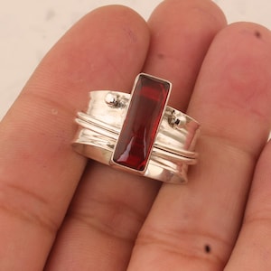 Könnte beinhalten: Ein silberner Ring mit einem roten Edelstein in rechteckiger Form. Der Ring hat ein drehendes Banddesign.