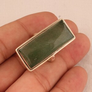 May include: A rectangular ring with a silver-colored band and a large, green stone. The stone has a slightly speckled appearance and is set within a silver frame. The ring is shown against a neutral background.