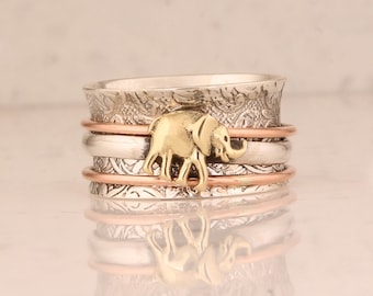 Puede incluir: Un anillo giratorio de plata con un dije de elefante de oro. El anillo tiene tres bandas, una de cobre, una de plata y una de plata con un diseño floral.
