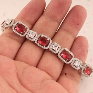 May include: A silver bracelet featuring alternating red and white gemstones. The red stones are oval-shaped, while the white stones are square. Each stone is set in a square frame and surrounded by smaller, clear stones.