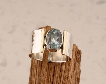 May include: A silver ring with a hammered texture band and gold accents. The ring features a large, oval-shaped, light blue gemstone. The ring is displayed on a piece of wood.