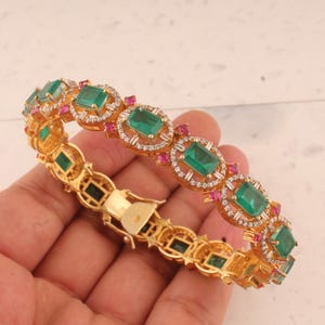 May include: Gold-toned bracelet featuring emerald-cut green gemstones, small red gemstones, and clear accent stones. The bracelet has a hinged clasp and a repeating pattern of gemstones. The bracelet is a piece of jewelry.